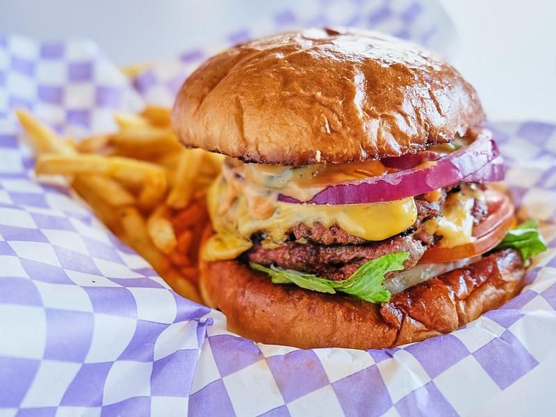 Doble smash burger con queso derretido, cebolla morada y papas sobre papel de cuadros morados.